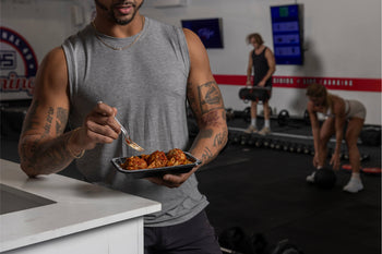 Man in gym eating a meal with meatballs and pasta, with others working out in the background.