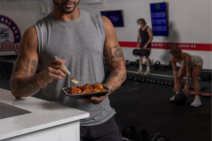 Man in gym eating a meal with meatballs and pasta, with others working out in the background.